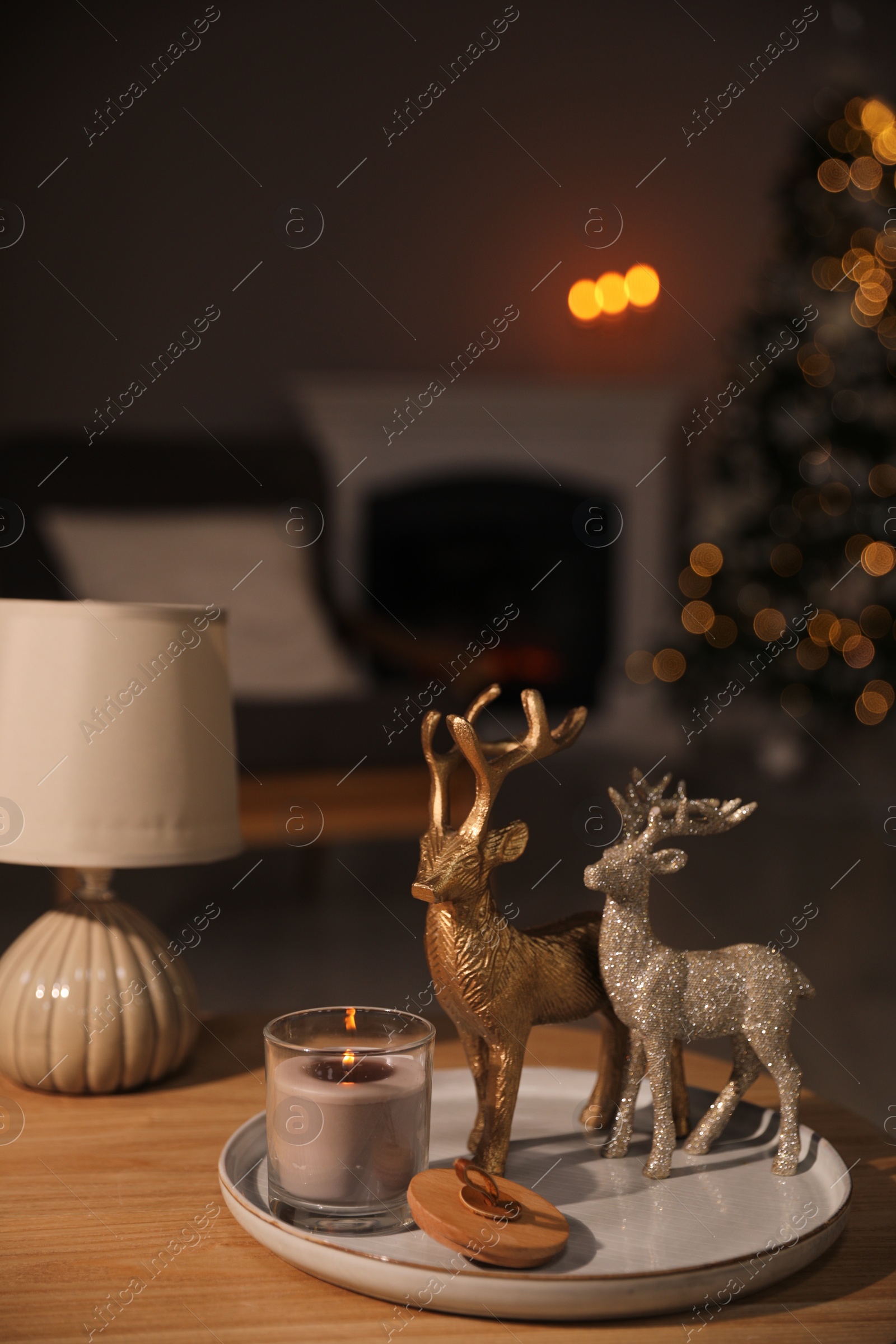Photo of Deer figures, burning candle with lamp on wooden table and Christmas tree in living room