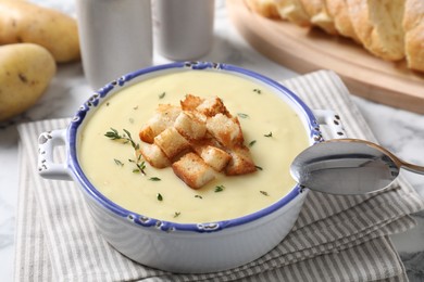 Photo of Tasty potato soup with croutons in ceramic pot and spoon on white table