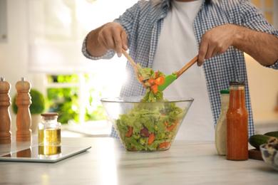 Photo of Man with tablet cooking salad at table in kitchen, closeup