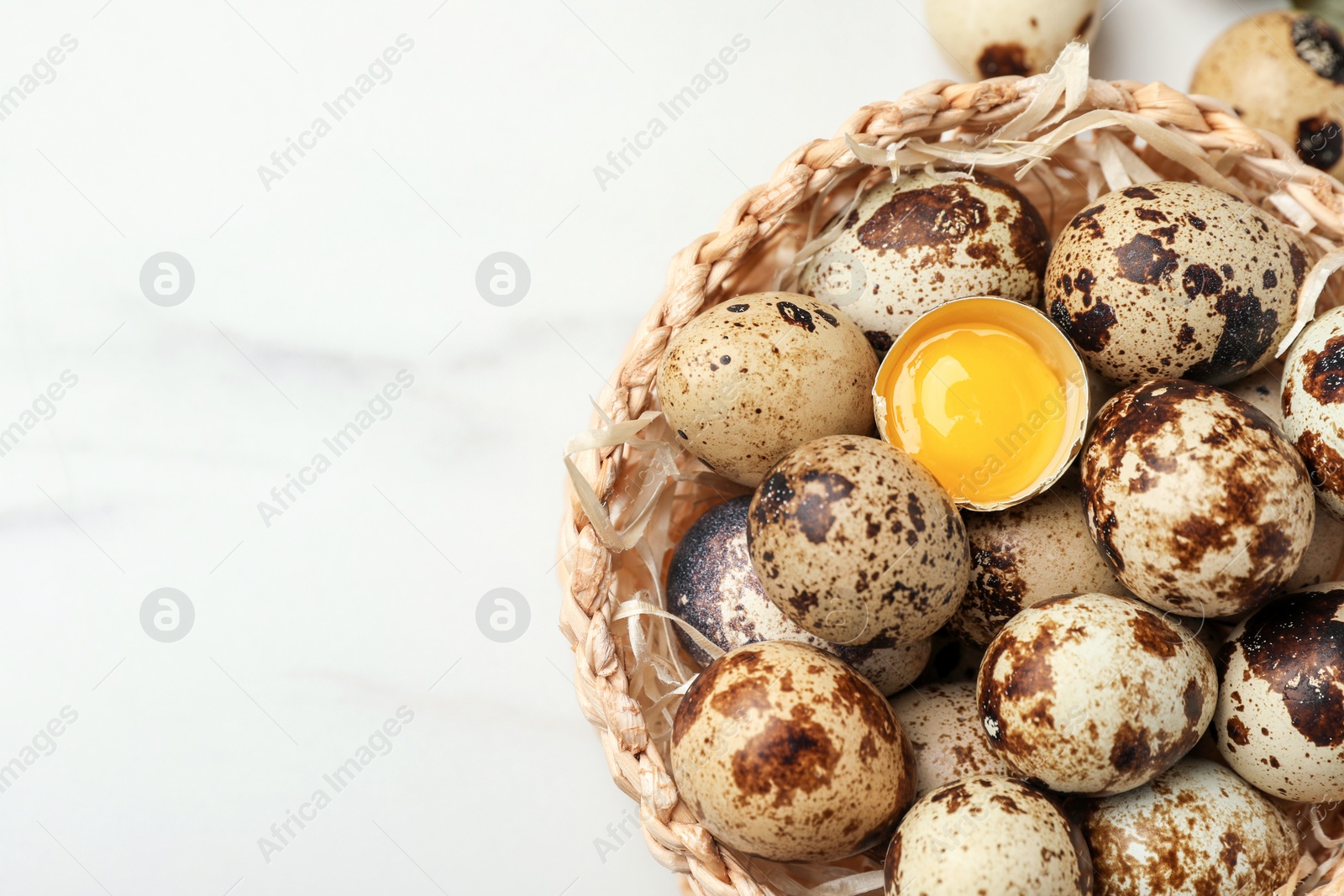 Photo of Wicker bowl with whole and cracked quail eggs on white marble table, top view. Space for text