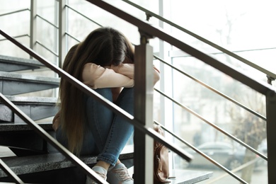 Upset teenage girl with backpack sitting on stairs indoors. Space for text