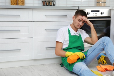 Exhausted janitor sitting on floor in kitchen, space for text. Cleaning service