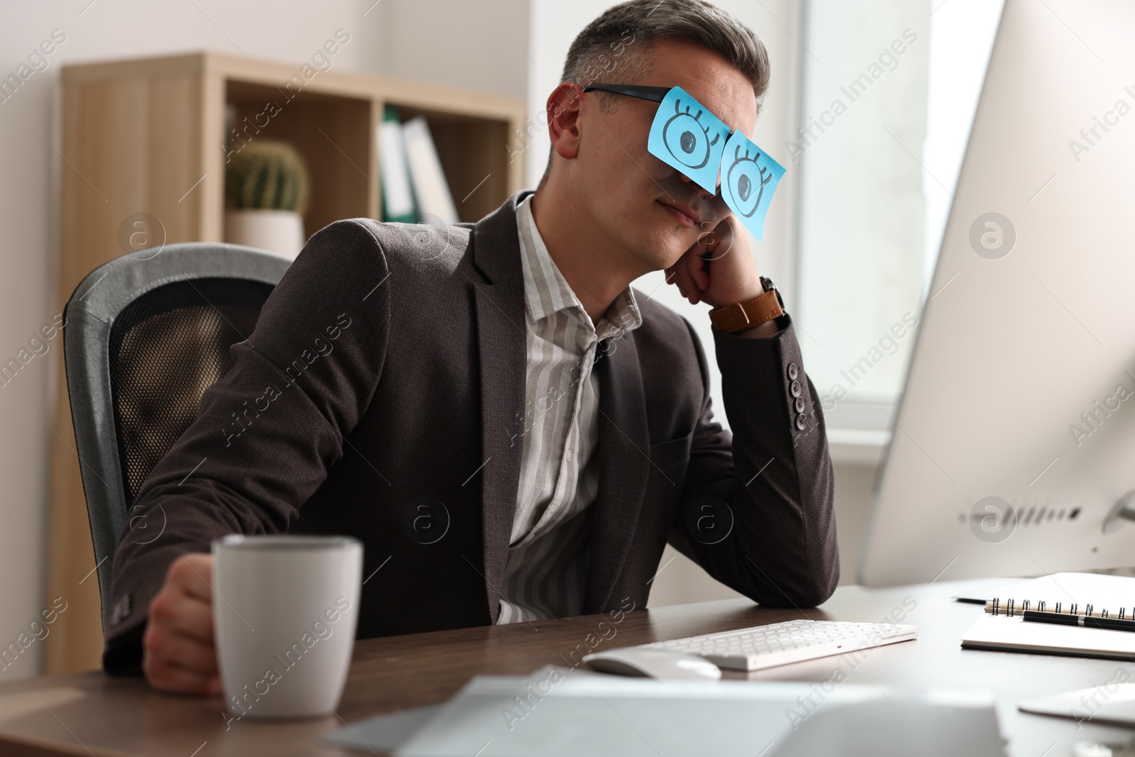 Photo of Man with fake eyes painted on sticky notes snoozing at workplace in office