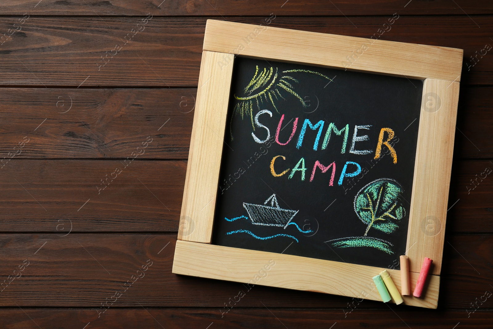 Photo of Small blackboard with text SUMMER CAMP, drawings and chalk sticks on wooden background, top view. Space for design