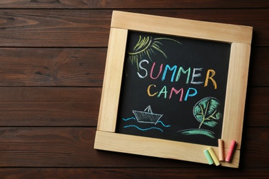 Photo of Small blackboard with text SUMMER CAMP, drawings and chalk sticks on wooden background, top view. Space for design