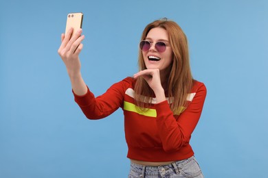 Beautiful woman in sunglasses taking selfie on light blue background