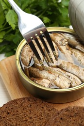 Photo of Fork with canned sprats and bread on wooden board, closeup
