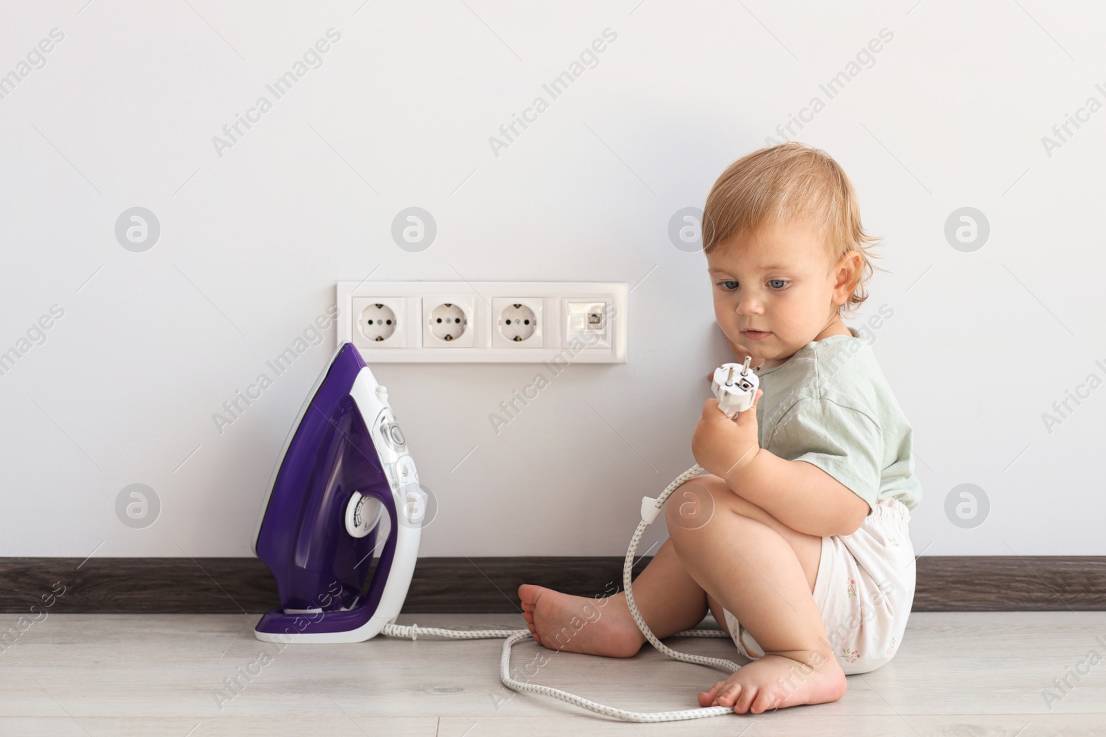 Photo of Cute baby playing with electrical socket and iron plug at home. Dangerous situation