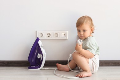 Photo of Cute baby playing with electrical socket and iron plug at home. Dangerous situation