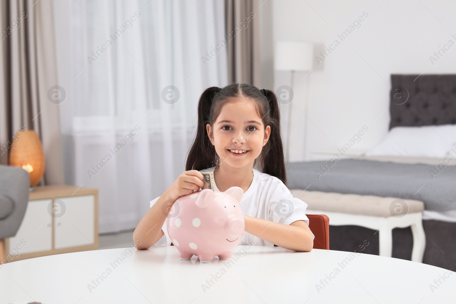 Photo of Little girl putting money into piggy bank at table indoors