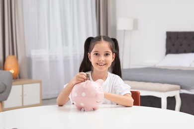 Photo of Little girl putting money into piggy bank at table indoors