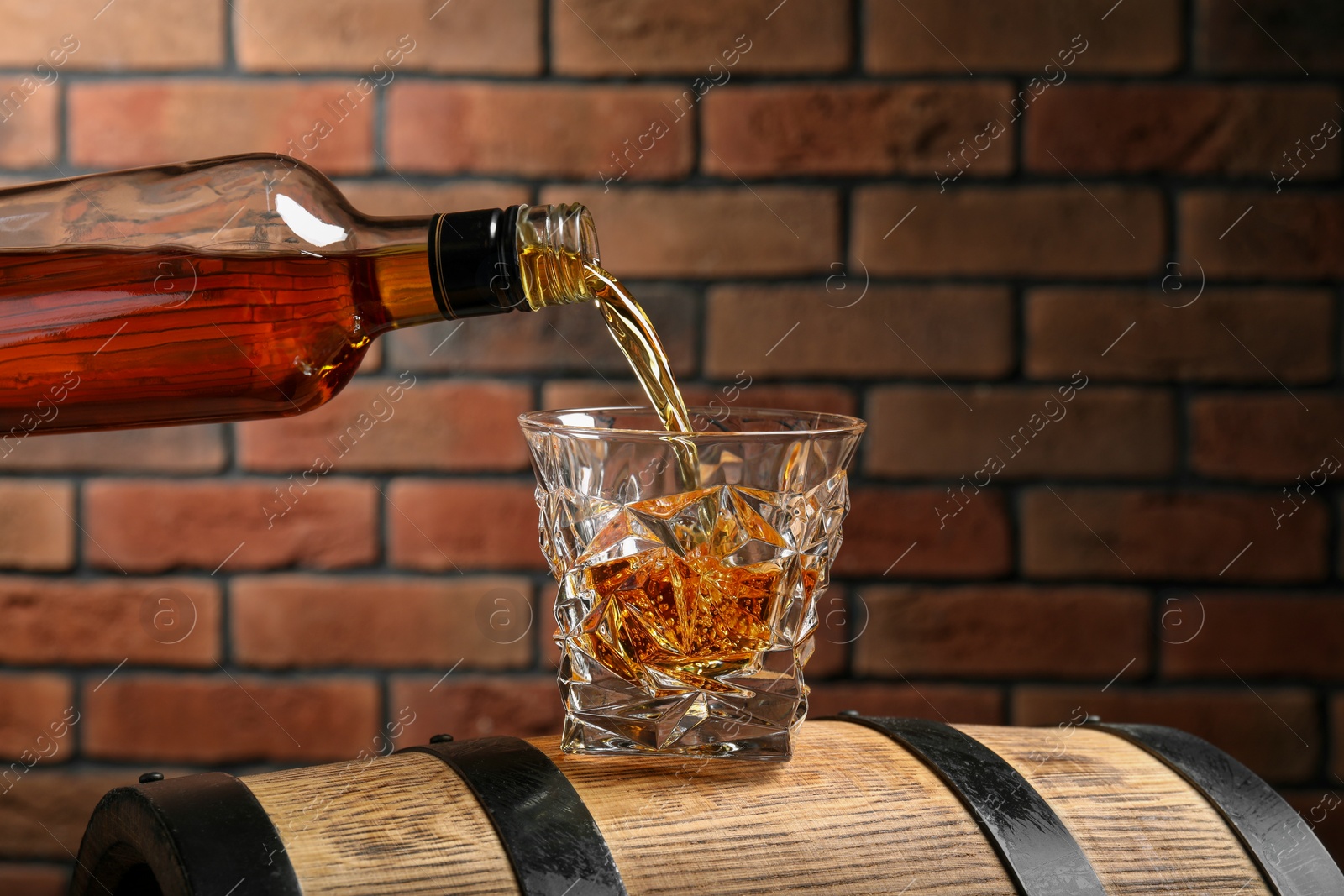 Photo of Pouring whiskey from bottle into glass on wooden barrel against brick wall