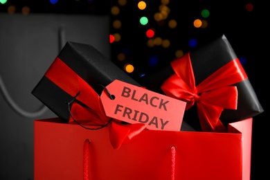 Photo of Paper shopping bags, gift boxes and tag with text Black friday against blurred lights, closeup