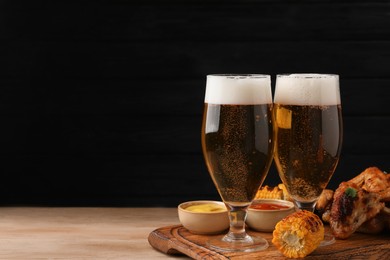 Delicious baked chicken wings, grilled corn and glasses with beer on wooden table against black background. Space for text