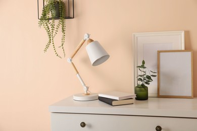 Photo of Stylish modern desk lamp, books and plant on white chest of drawers near beige wall indoors
