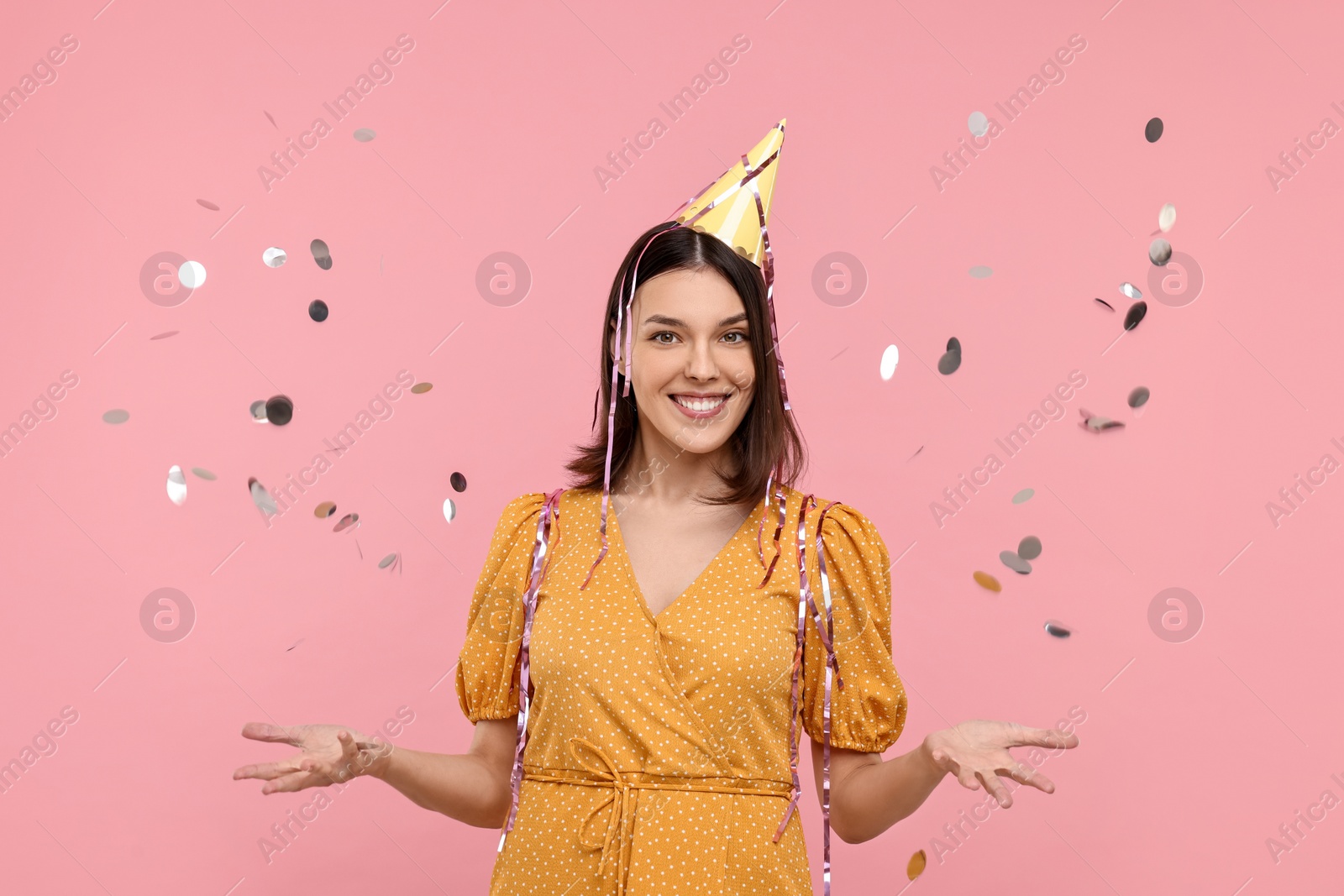 Photo of Happy young woman in party hat near flying confetti on pink background