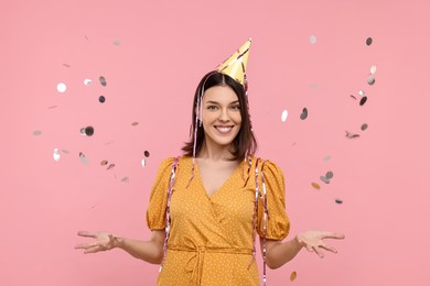 Happy young woman in party hat near flying confetti on pink background