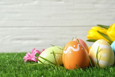 Photo of Colorful painted Easter eggs and flowers on green grass against wooden background, space for text