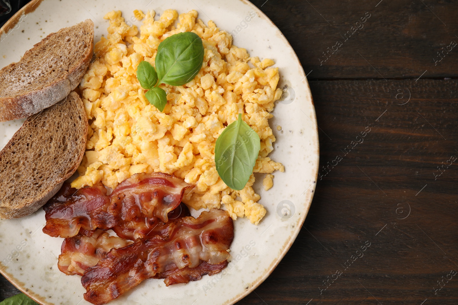 Photo of Delicious scrambled eggs with bacon and basil in plate on wooden table, top view. Space for text