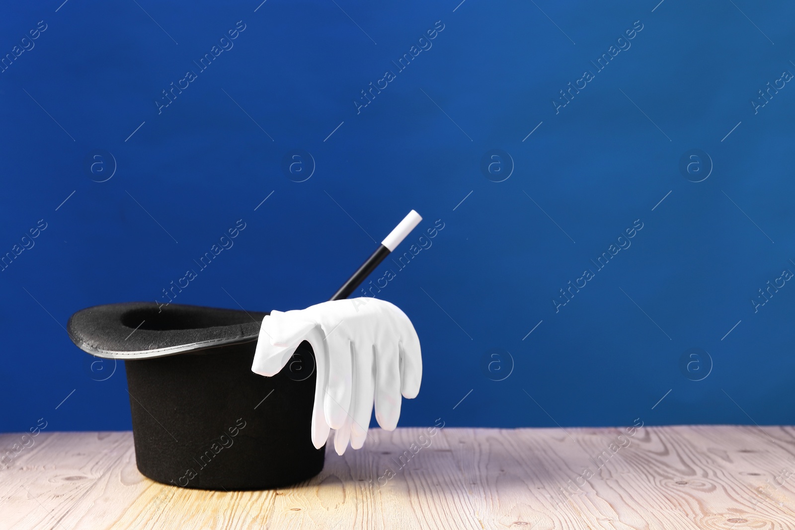 Photo of Magician's hat, wand and gloves on wooden table against blue background, space for text