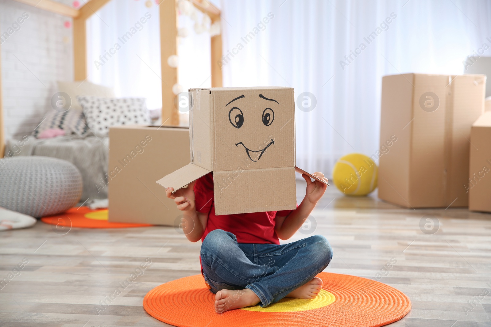 Photo of Cute little child wearing cardboard box with smiling face in bedroom