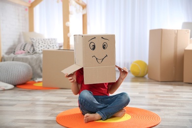 Photo of Cute little child wearing cardboard box with smiling face in bedroom