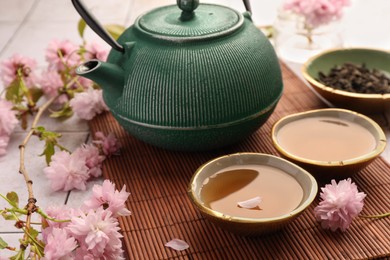 Photo of Traditional ceremony. Cup of brewed tea, teapot and sakura flowers on bamboo mat, closeup
