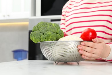 Photo of Woman holding colander with fresh vegetables at white marble table in kitchen, closeup