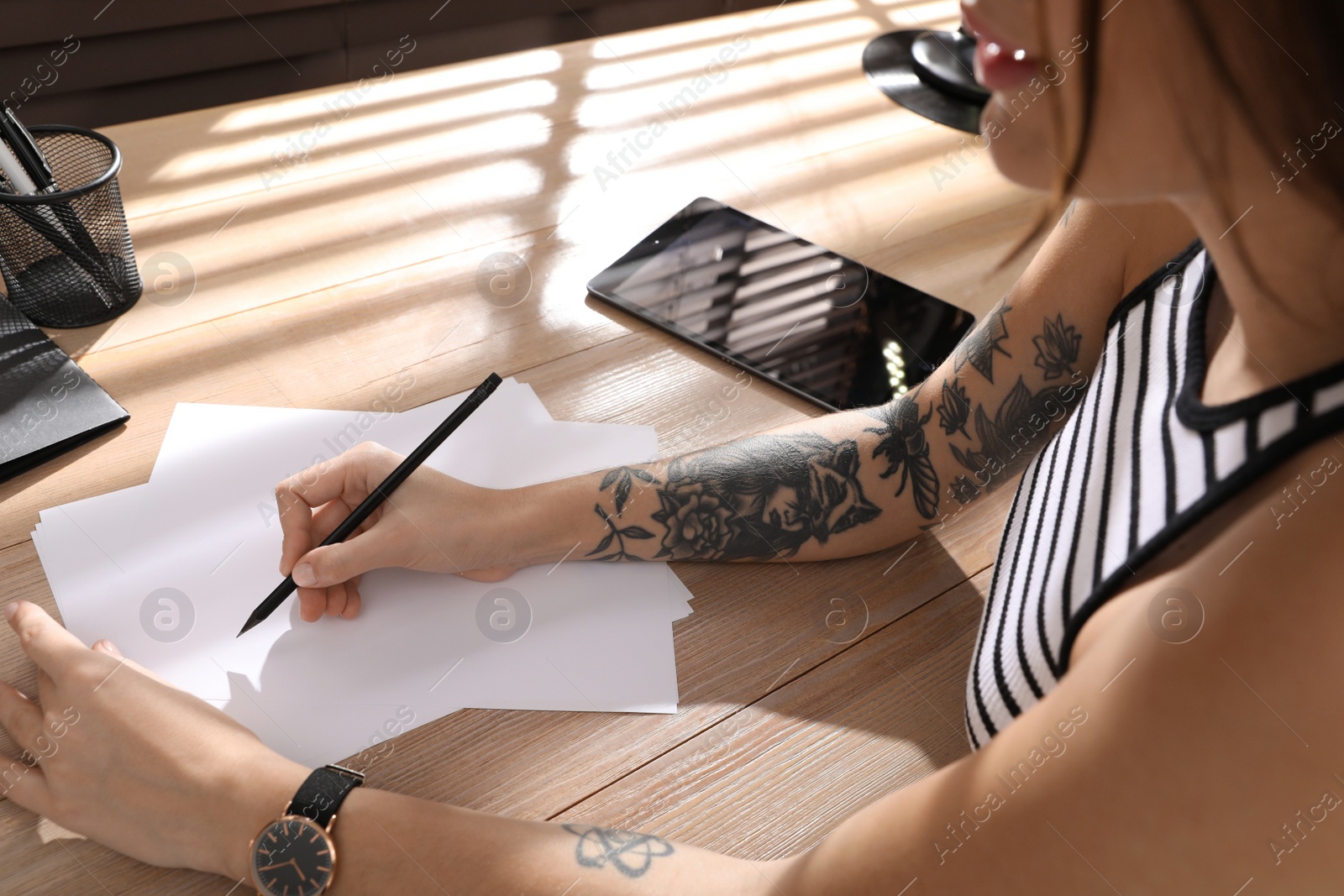 Photo of Beautiful woman with tattoos on arms drawing in sketchbook at table, closeup