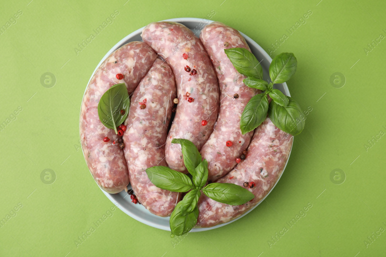 Photo of Raw homemade sausages, basil leaves and peppercorns on green table, top view