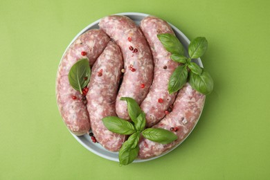 Photo of Raw homemade sausages, basil leaves and peppercorns on green table, top view