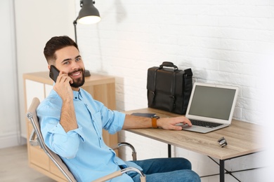 Handsome young man talking on phone while working at table with laptop in home office