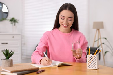 Photo of Young woman with glass of water writing in notebook at wooden table indoors