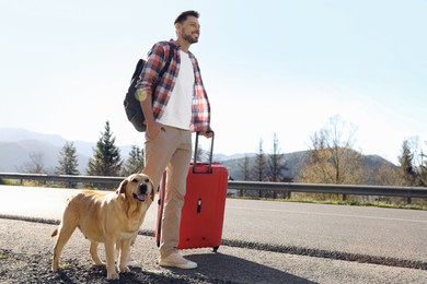 Photo of Happy man with red suitcase and adorable dog near road, space for text. Traveling with pet