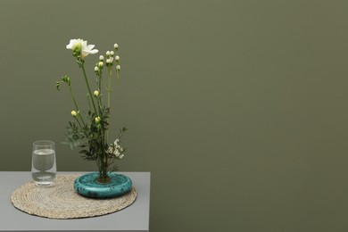 Photo of Stylish ikebana with beautiful flowers, green branches and glass of water on gray table near olive wall, space for text