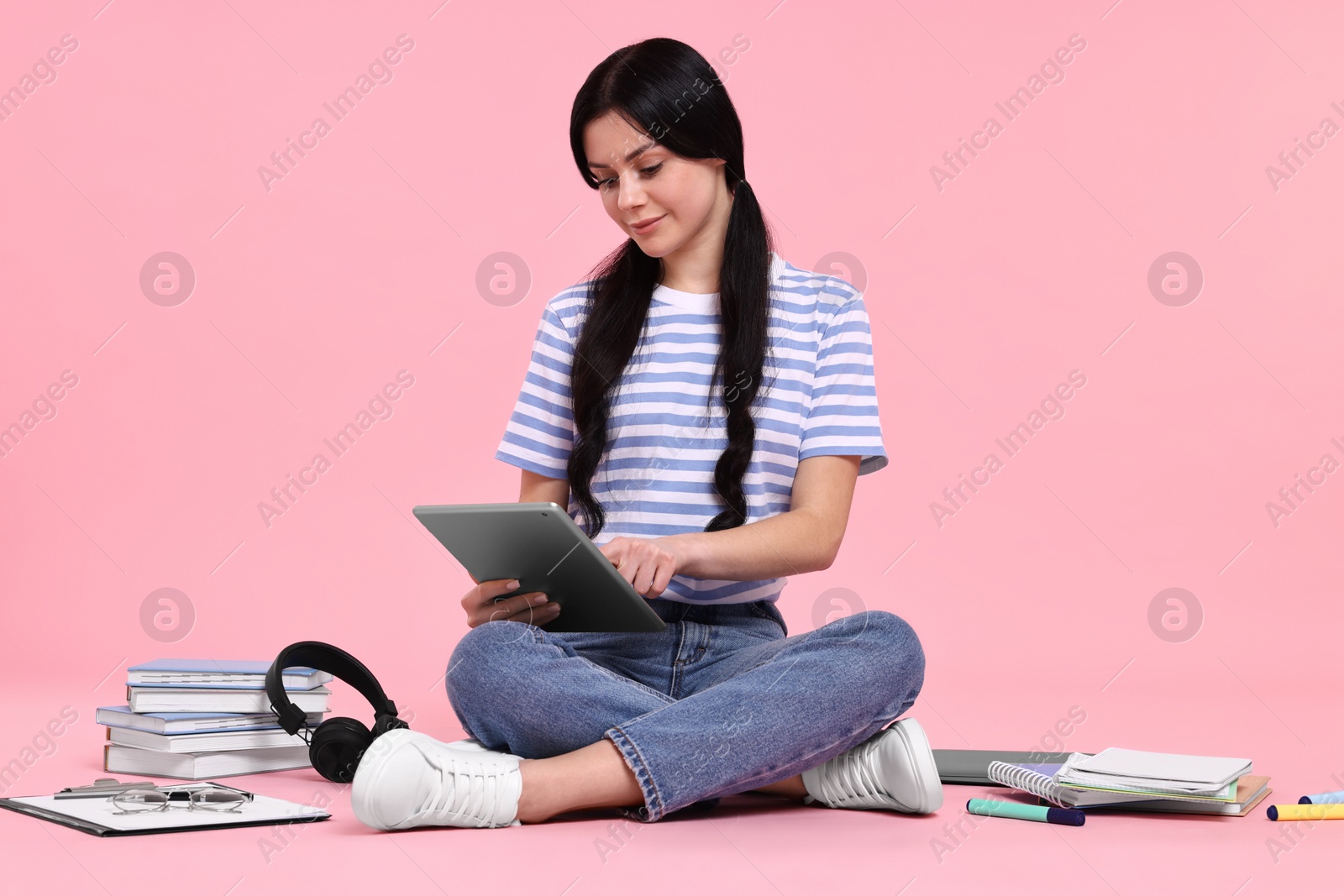 Photo of Student with tablet sitting among books and stationery on pink background