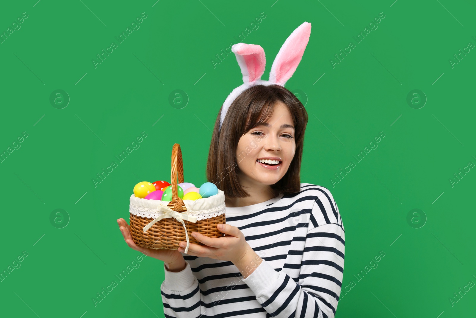 Photo of Easter celebration. Happy woman with bunny ears and wicker basket full of painted eggs on green background