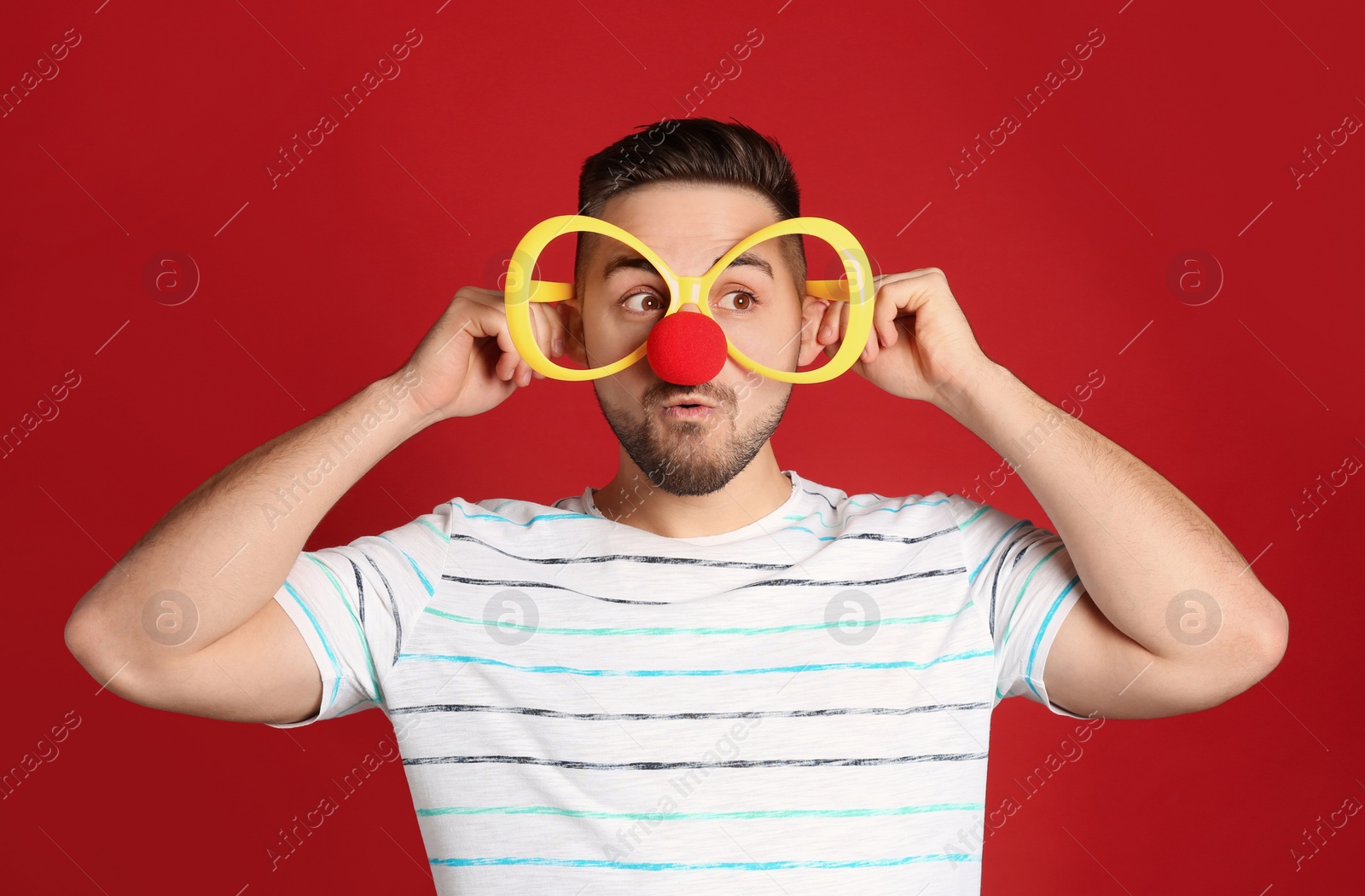 Photo of Emotional young man with party glasses and clown nose on red background. April fool's day