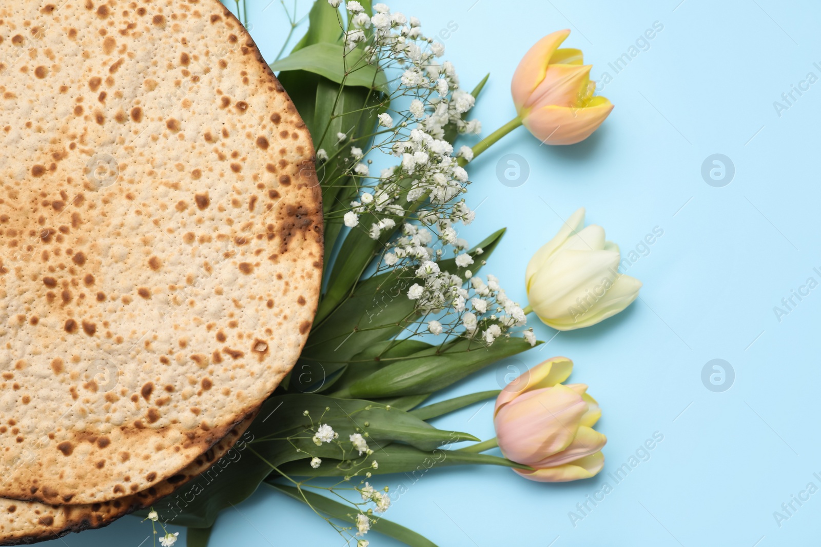 Photo of Tasty matzos and fresh flowers on light blue background, flat lay. Passover (Pesach) celebration