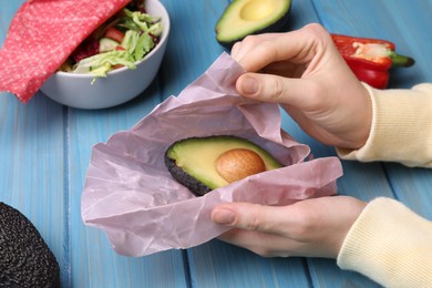 Photo of Woman packing half of fresh avocado into beeswax food wrap at light blue wooden table, closeup