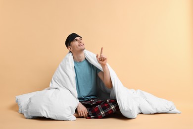 Photo of Happy man in pyjama and sleep mask wrapped in blanket on beige background