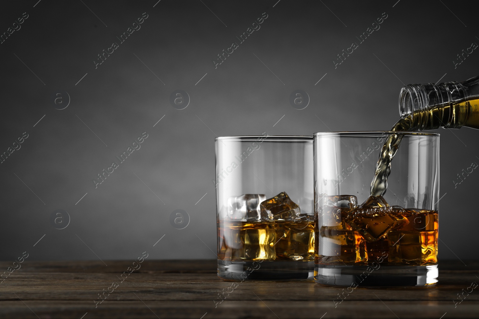 Photo of Pouring whiskey into glass with ice cubes at wooden table against grey background, space for text