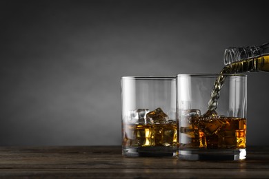 Photo of Pouring whiskey into glass with ice cubes at wooden table against grey background, space for text