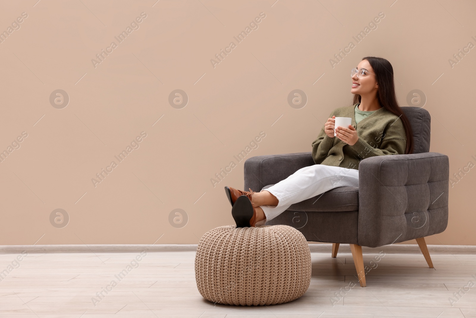 Photo of Beautiful woman with cup of drink sitting in armchair near beige wall indoors, space for text