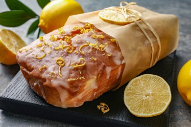 Photo of Wrapped tasty lemon cake with glaze and citrus fruits on grey table, closeup