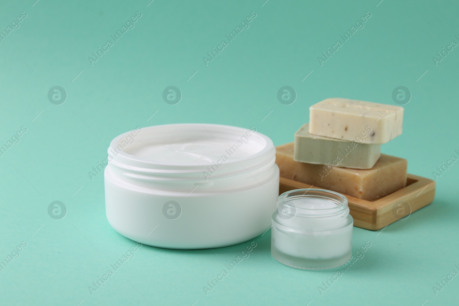 Photo of Moisturizing cream in open jars and soap bars on turquoise background. Body care products