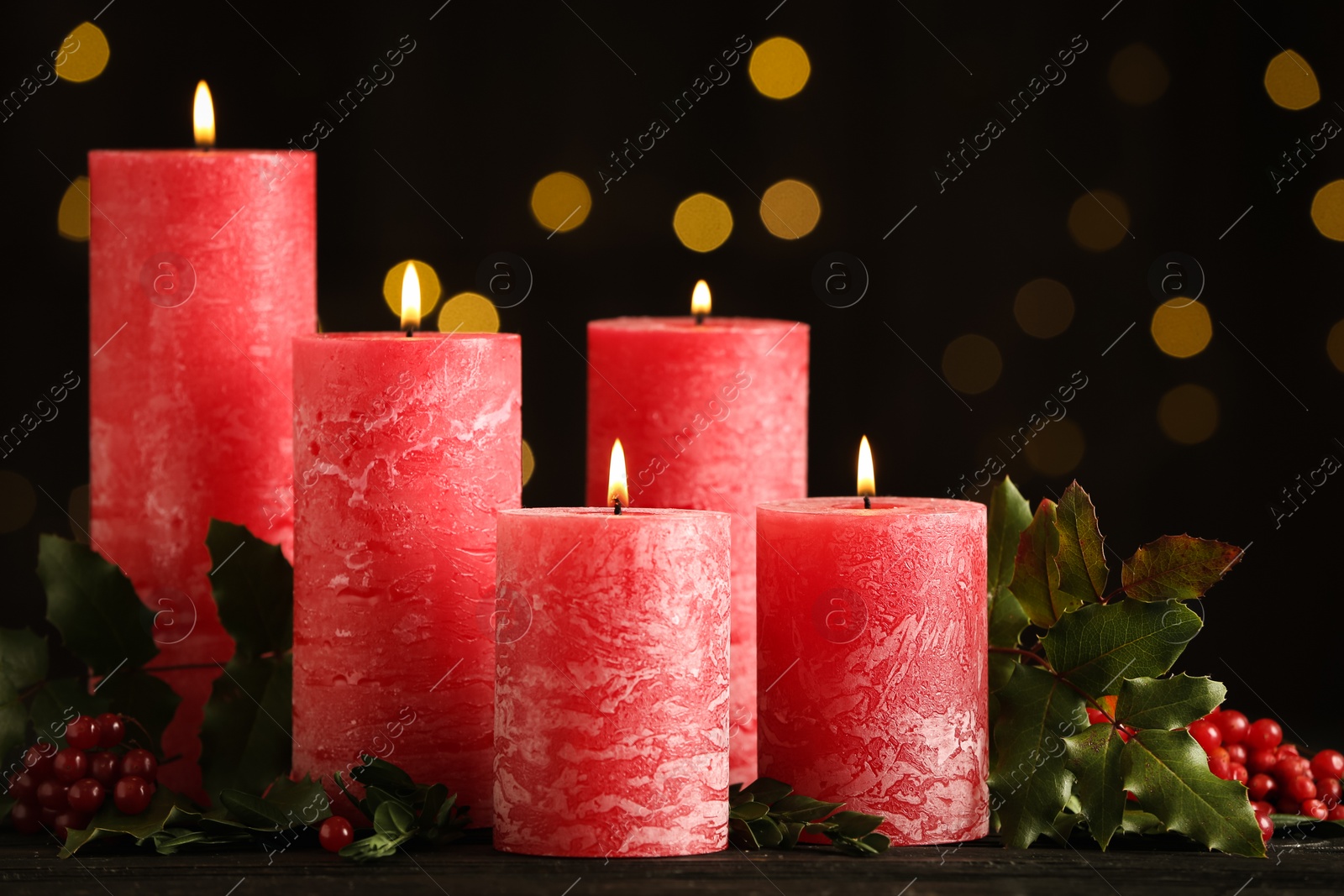 Photo of Burning red candles with holly branches on table against blurred Christmas lights
