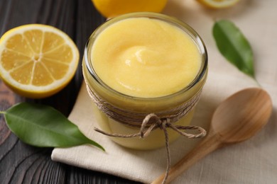 Photo of Delicious lemon curd in bowl, fresh citrus fruit, spoon and green leaves on wooden table, closeup