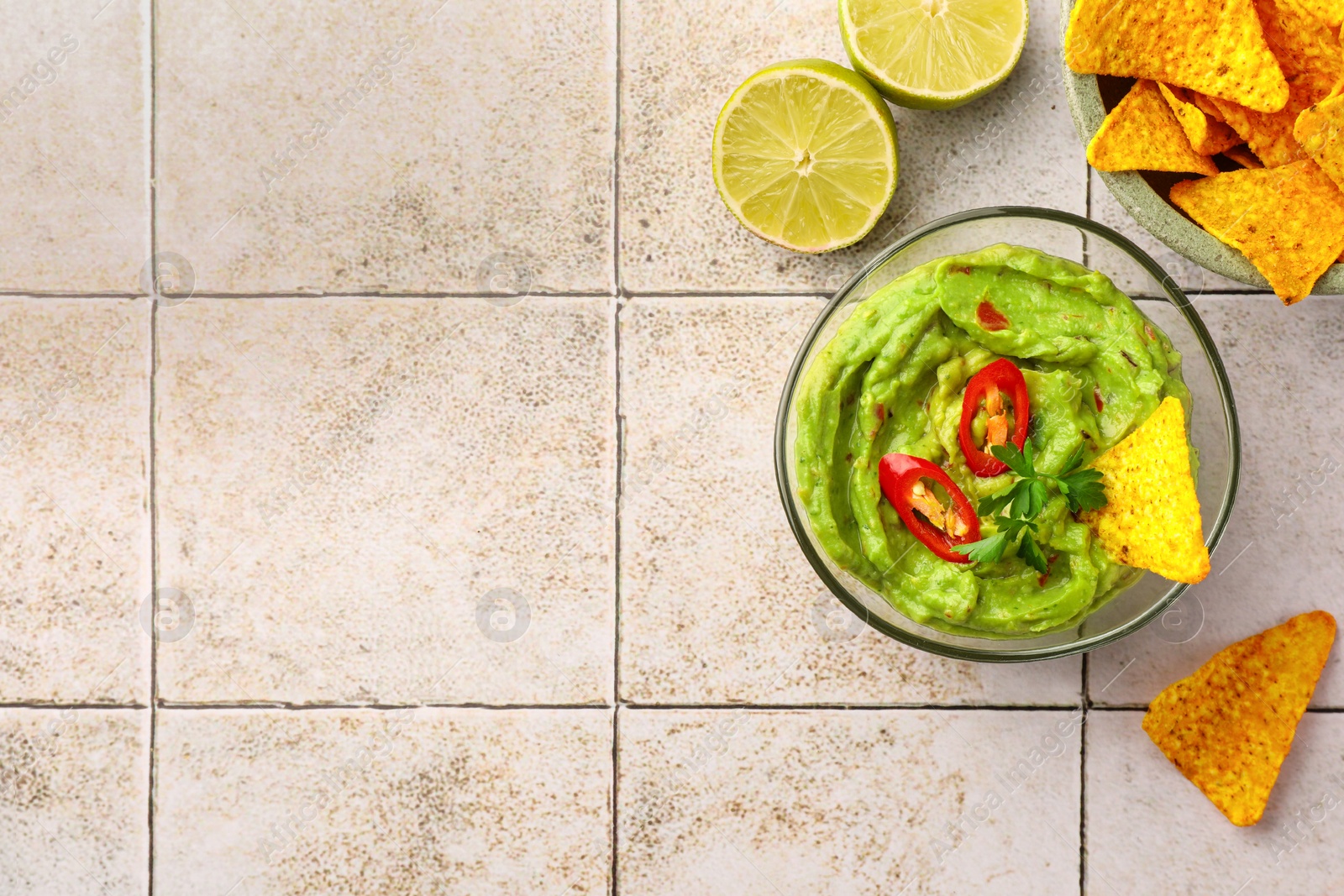 Photo of Bowl of delicious guacamole with chili pepper, nachos chips and lime on white tiled table, flat lay. Space for text