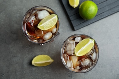Photo of Glasses of cocktail with cola, ice and cut lime on grey background, flat lay